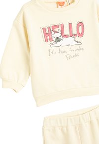 Ljusgul sweatshirt och byxset med en grafisk bild av en hund med texten "HEJ" och "Dags att skaffa vänner," ribbade muddar.