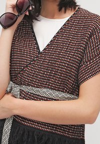 Svart och orange vävd wrap med ett geometriskt mönster, som har ett vitt flätat bälte och texturerad frans i fållen.