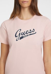 Jasnoróżowa koszulka bawełniana z haftowanym, granatowym logo "Guess Jeans 1981" w stylu kursywy. Krótkie rękawy i okrągły dekolt. Miękka tekstura.