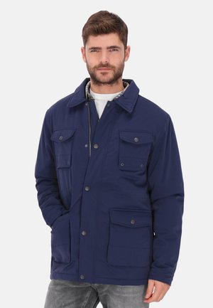 Mand iført en marineblå jakke med flere lukkede lommer over en hvid skjorte og grå jeans mod en ensfarvet lys baggrund.