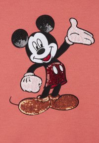 Topolino con pantaloni rossi e scarpe marroni di paillettes, sorridente con una mano alzata su uno sfondo di stoffa rosa.