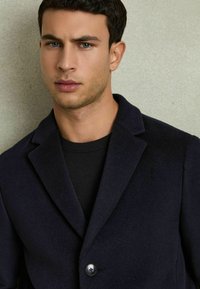 Marineblå uldblanding blazer med spidse lapeller, enkelt knaplukning og en tekstureret overflade, båret over en sort skjorte.