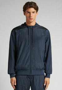 Marinblå jacka med huva, tillverkad av ett texturerat, glänsande material och elastiska ärmslut. Frontal dragkedja och sidofickor fulländar designen.