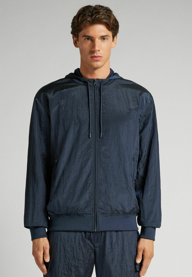 Marinblå jacka med huva, tillverkad av ett texturerat, glänsande material och elastiska ärmslut. Frontal dragkedja och sidofickor fulländar designen.