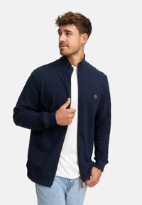 Joven vestido con una chaqueta navy con cremallera sobre una camisa blanca y jeans de color azul claro, mirando hacia la izquierda contra un fondo liso.