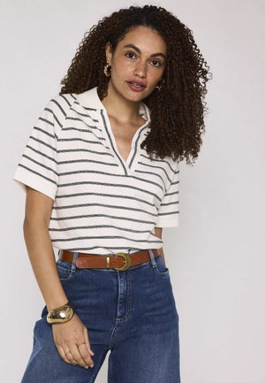 Femme aux cheveux bouclés portant un polo blanc et noir à rayures, un jean bleu, une ceinture marron, un bracelet en or et des boucles d'oreilles créoles.