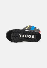 Suela de goma negra con un profundo grabado, con el logotipo "SOREL". La parte superior incluye correas coloridas en azul, amarillo y rojo sobre una base gris.