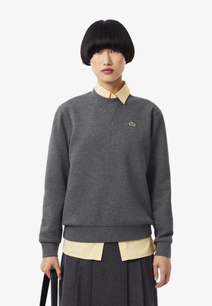 Persona con un corte de pelo bob negro corto, vistiendo un sudadera gris sobre una camisa amarilla de cuello y una falda plisada oscura, sosteniendo un bolso negro.