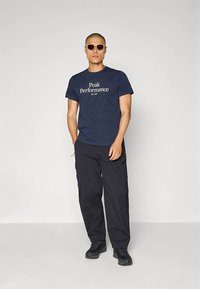 Navyblauw T-shirt met het logo van "Peak Performance", gecombineerd met zwarte cargobroeken en een zonnebril, gedragen door een persoon die rechtop staat.