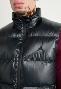 Man i svart glansig pufferväst med dragkedja och liten Jumpman-logotyp på vänster bröst över en mörkröd tröja.