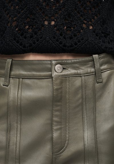 Κοντινό της μέσης με μαύρο πλεκτό τοπ και ψηλόμεσο χακί δ ερμάτινο παντελόνι με κουμπί και θηλιές για ζώνη.