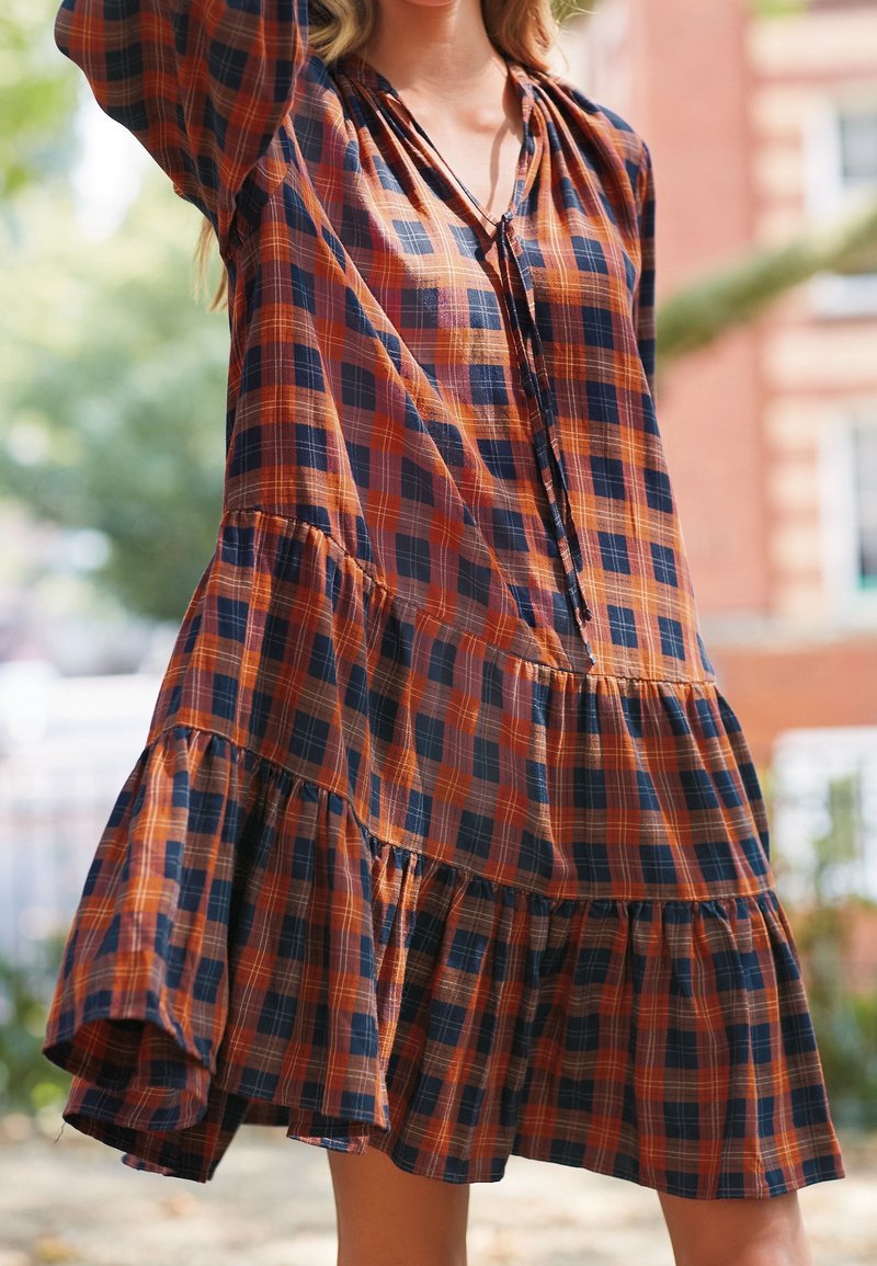 Femme portant une robe ample à étages avec un motif à carreaux orange et bleu marine, en extérieur avec des arbres et un bâtiment flous en arrière-plan.
