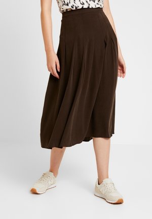 Femme portant une jupe midi marron foncé et des baskets blanches, debout avec les mains détendues le long du corps sur un fond uni.