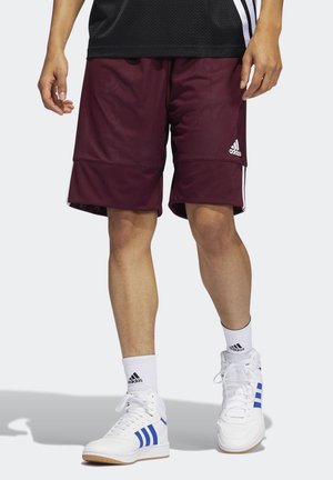 Personne portant un short de basket Adidas bordeaux, des chaussettes Adidas blanches et des baskets blanches à rayures bleues, debout sur un fond clair.