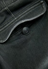 Tejido de mezclilla negro con una solapa de bolsillo cosida, que presenta un botón negro redondo con texto en relieve. Textura suave y un brillo sutil.