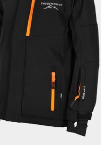 Veste noire avec des zips et des accents orange, comportant un logo sur la poitrine, plusieurs poches et des poignets ajustables pour un ajustement sécurisé.
