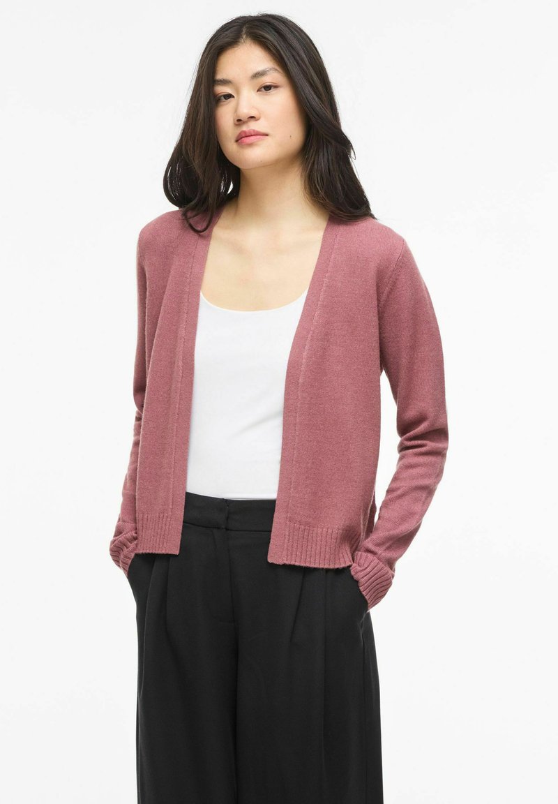 Mujer joven con cabello largo y oscuro, vistiendo un cárdigan abierto de color rosa polvoriento, camiseta blanca y pantalones negros de pierna ancha, de pie con las manos en los bolsillos.