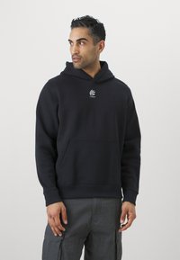Svart hettegenser med frontlomme, ribbede mansjetter og en liten hvit logo på brystet. Stoffet ser mykt og texturert ut.