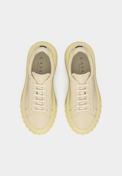 Ζευγάρι εκρού δερμάτινα αθλητικά παπούτσια με παχιές κίτρινες σόλες και λευκά κορδόνια, σε πλάγια θέα από πάνω σε απλό φόντο.