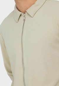 Light beige zip-up jacket with a collared neckline, ribbed texture, and metal zipper. Features long sleeves and a fitted silhouette.