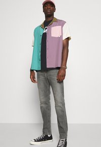 Multicolored short-sleeve shirt featuring green, purple, cream, and pink panels, with a collar and a front pocket. Worn with gray jeans and black sneakers.
