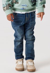 Blå denimbyxor med dragsnodd i midjan. Barnet har på sig gräddfärgade sneakers med kardborreband och vita snören.