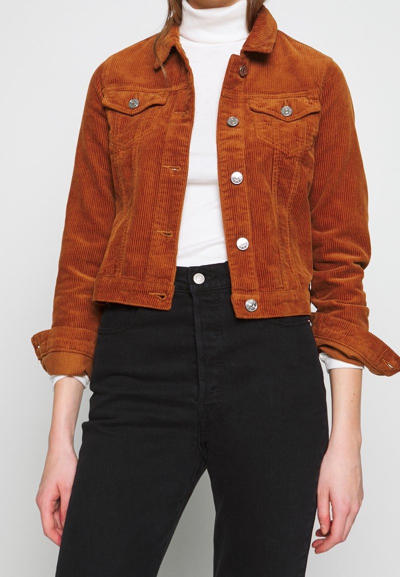 Vrouw in een roestkleurige corduroy jas over een witte coltrui en hoog-getailleerde zwarte jeans, met opgerolde mouwen van de jas.