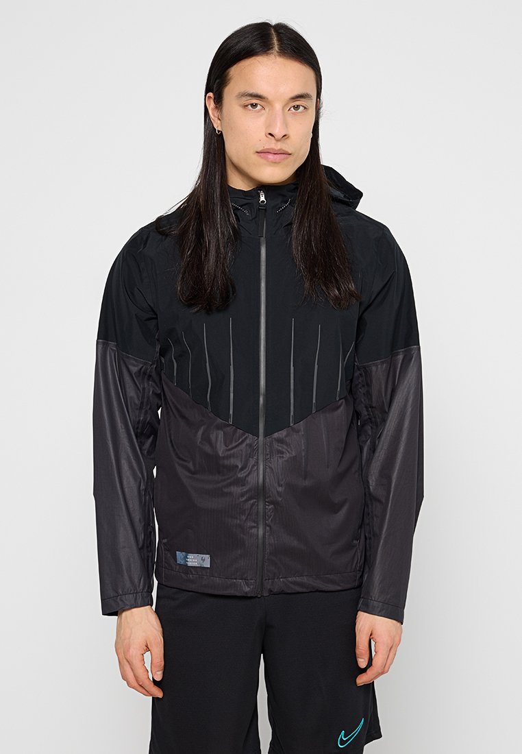 Zwarte sportjack met capuchon, met contrasterende texturen en verticale ontwerplijnen op de borst. Ritsluiting en logo-detail zichtbaar.