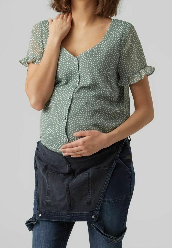 Hellgrüne Bluse mit weißen Punkten und kurzen, gerafften Ärmeln, kombiniert mit einer dunklen Schürzen-ähnlichen Denim-Blende über blauen Jeans, die Passform und Komfort verbessert.