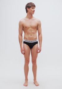 Μαύρα μποξεράκια με λευκή μέση που φέρει το κείμενο "Sounds gay I'm in." Το μοντέλο στέκεται ξυπόλητο μπροστά σε μια απλή, μονόχρωμη επιφάνεια.