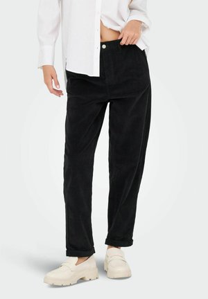 Personne portant un pantalon noir ample remonté aux chevilles, une chemise blanche et des mocassins épais couleur blanc cassé, debout devant un fond uni.