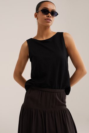 Donna che indossa un top nero senza maniche, gonna scura, occhiali da sole ovali neri e orecchini a perno in argento, posando con le mani dietro la schiena.