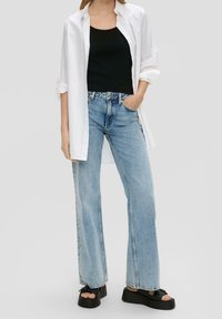 Svart linne under en vit skjorta med knappar, kombinerat med ljusblå utsvängda jeans och svarta platåsandaler. Slät textur genom hela outfiten.