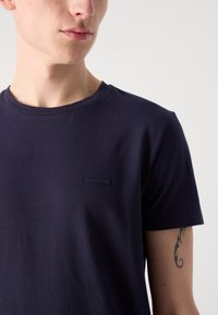 Tamsiai mėlynas medvilnės marškinėliai su apvalia iškirpte, su maža tekstūruota Calvin Klein etikete kairiajame krūtinės srityje.