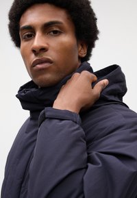 Giacca puffer blu navy con colletto alto, caratterizzata da un tessuto texturizzato e chiusura con zip. La mano del modello sistema il colletto, mettendo in risalto il design elegante.