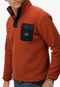 Orange fleece pullover with high collar, black chest pocket, and button details. Soft texture, casual fit, and mountain logo on the pocket.