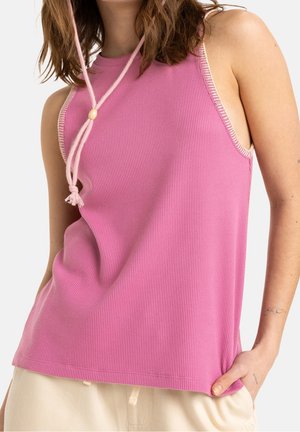 Kvinde iført en ærmeløs lyserød ribstrikket tanktop med cremeforet kant og lysebeige bukser, der viser en afslappet positur med den ene hånd i lommen.