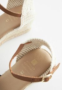 Par de sandalias de cuña tejidas en beige con correas de tobillo de cuero marrón y hebillas doradas sobre fondo blanco.