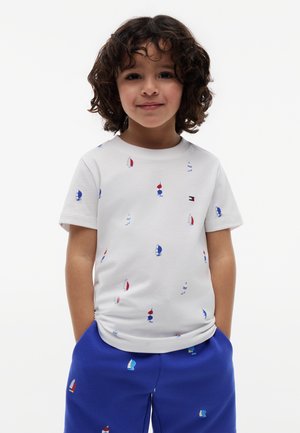 Barn med krøllet hår står med hænderne i lommerne, iført en hvid t-shirt og blå shorts med små farverige sejlbåde.