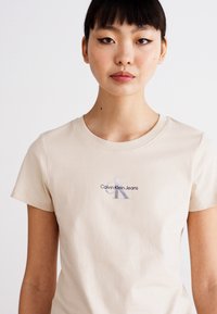 Bežové bavlnené tričko s okrúhlym výstrihom, s reflexným logom "Calvin Klein Jeans" a stylizovaným "K" na hrudi. Krátke rukávy.