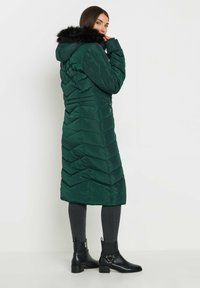 Cappotto imbottito verde lungo con motivo a chevron trapuntato, cappuccio foderato in pelliccia e tasche laterali. Abbinato a stivali neri alla caviglia e jeans scuri.