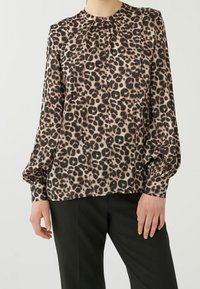 Blouse à manches longues imprimé léopard en tissu léger, avec un col froncé, une patte de boutonnage et des poignets élastiques. Portée avec un pantalon noir.