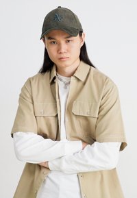 Lys beige skjorte med korte ermer og to lommer foran, lagd over en hvit langermet skjorte. Grønn cap med brodert logo. Nøytral bakgrunn.