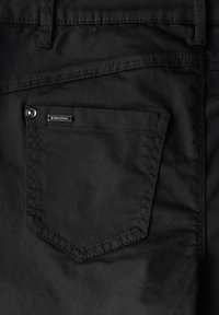 Pantalones negros con un bolsillo trasero que tiene una solapa redondeada, detalles de costura sutiles y un pequeño acento de logotipo metálico cerca del bolsillo.