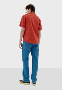 Kurzärmliges, orange-rotes Shirt aus strukturiertem Stoff, kombiniert mit lässigen blauen Jeans. Bequeme Schuhe runden das Outfit ab.
