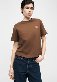 Νεαρή γυναίκα που φοράει καφέ κοντομάνικη μπλούζα με μικρό λογότυπο στο στήθος και σκούρο μπλε τζιν, ποζάροντας με το ένα χέρι στην τσέπη.