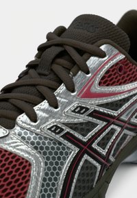 Asics futócipő ezüst hálós felsőrésszel, barna fűzőkkel és piros díszítésekkel. Hatszögletű mintázatokkal és jól látható márkajelzéssel a nyelvén.