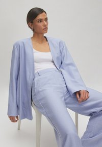 Lichtblauwe oversized blazer en bijpassende broek van textiel, gedragen over een witte tanktop, zittend op een stoel tegen een neutrale achtergrond.