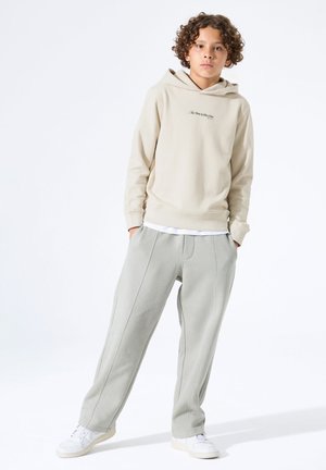 Beige hooded sweatshirt met logo aan de voorkant, gecombineerd met lichtgrijze, losvallende joggingbroek met zijnaden en zakken. Witte sneakers.