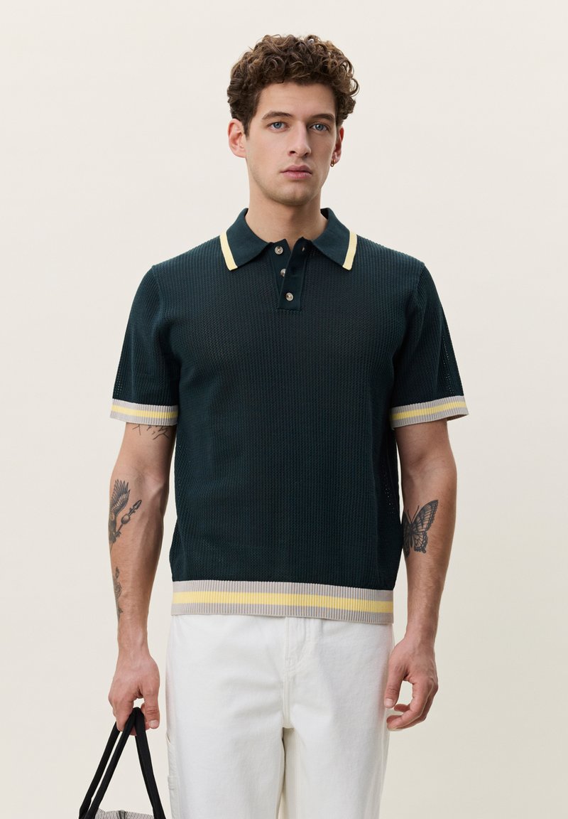 Jeune homme aux cheveux bouclés portant un polo en maille noire avec des rayures jaunes, un pantalon blanc, tenant un sac noir à poignée.